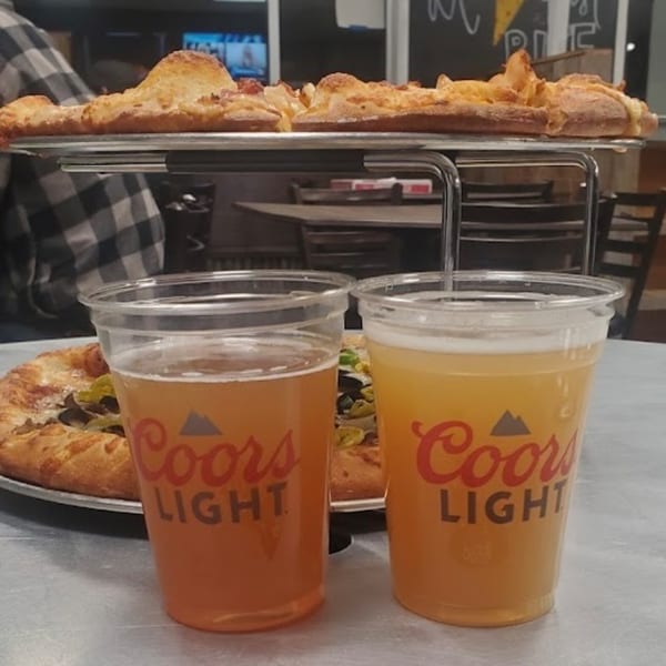 Two Coors Light beers and two pizzas, one on a stand and one on the table.